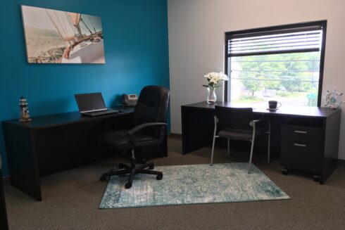 A modern office space featuring two black desks arranged in an L-shape, with one section against a bold blue wall and the other by a window overlooking a street.