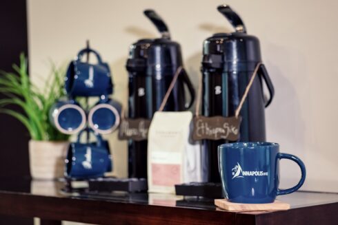 A coffee station featuring two black airpot coffee dispensers labeled "Mura" and "Ethiopia Sidamo," a bag of coffee beans, and a dark blue mug resting on a wooden coaster.