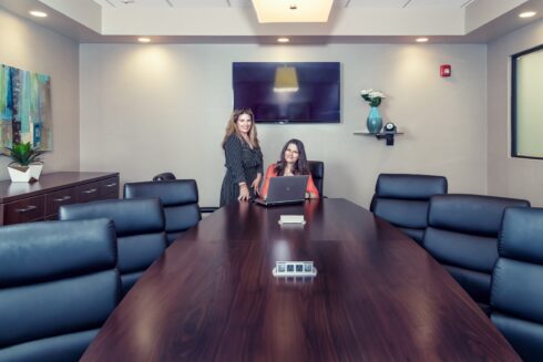 Two smiling women standing and seated at a large, dark wooden conference table surrounded by black leather chairs in a modern meeting room.