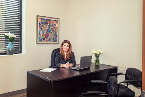 A smiling professional woman with long brown hair, wearing a striped blazer, seated at a dark executive desk with a laptop and flowers in a bright, modern office.