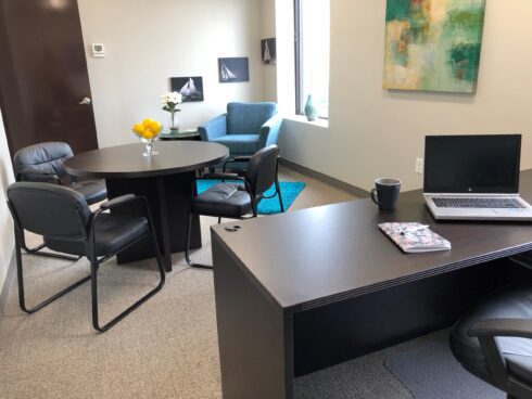 Office space with a large black executive desk in the foreground and a small meeting table and sitting area in the background, featuring nautical decor.
