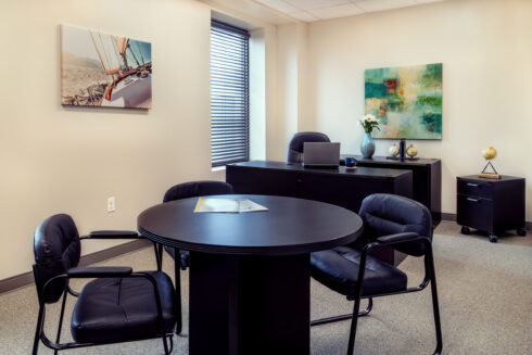 Office space with a dark black executive desk and a circular meeting table in the foreground, featuring nautical and abstract art decor.