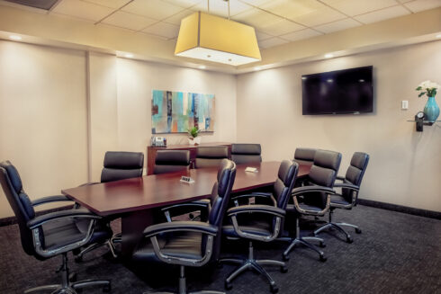 Empty, modern corporate conference room with a dark mahogany table, eight black leather executive chairs, and a flat-screen TV on the wall.