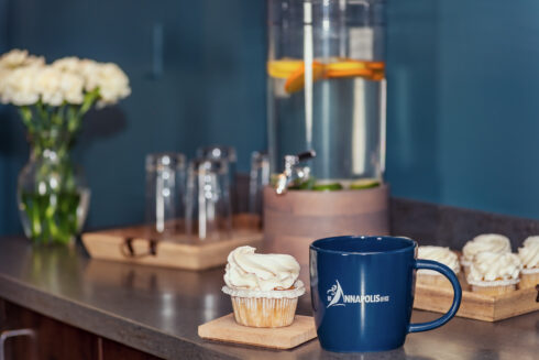 Close-up of a counter with a cupcake, a blue mug with a logo, a fruit-infused water dispenser, and a vase of white flowers.