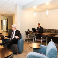 Two businessmen working on laptops in a modern, casual office lounge or waiting area with comfortable seating and small tables.