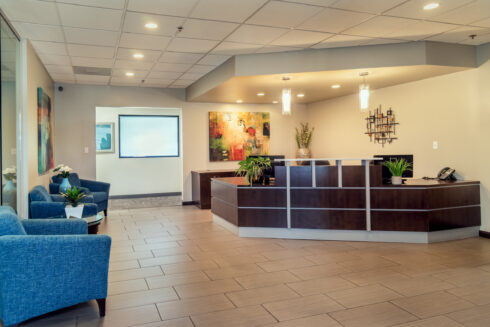 Wide shot of a modern, well-decorated office reception area with a dark wood front desk, blue seating, and abstract artwork.