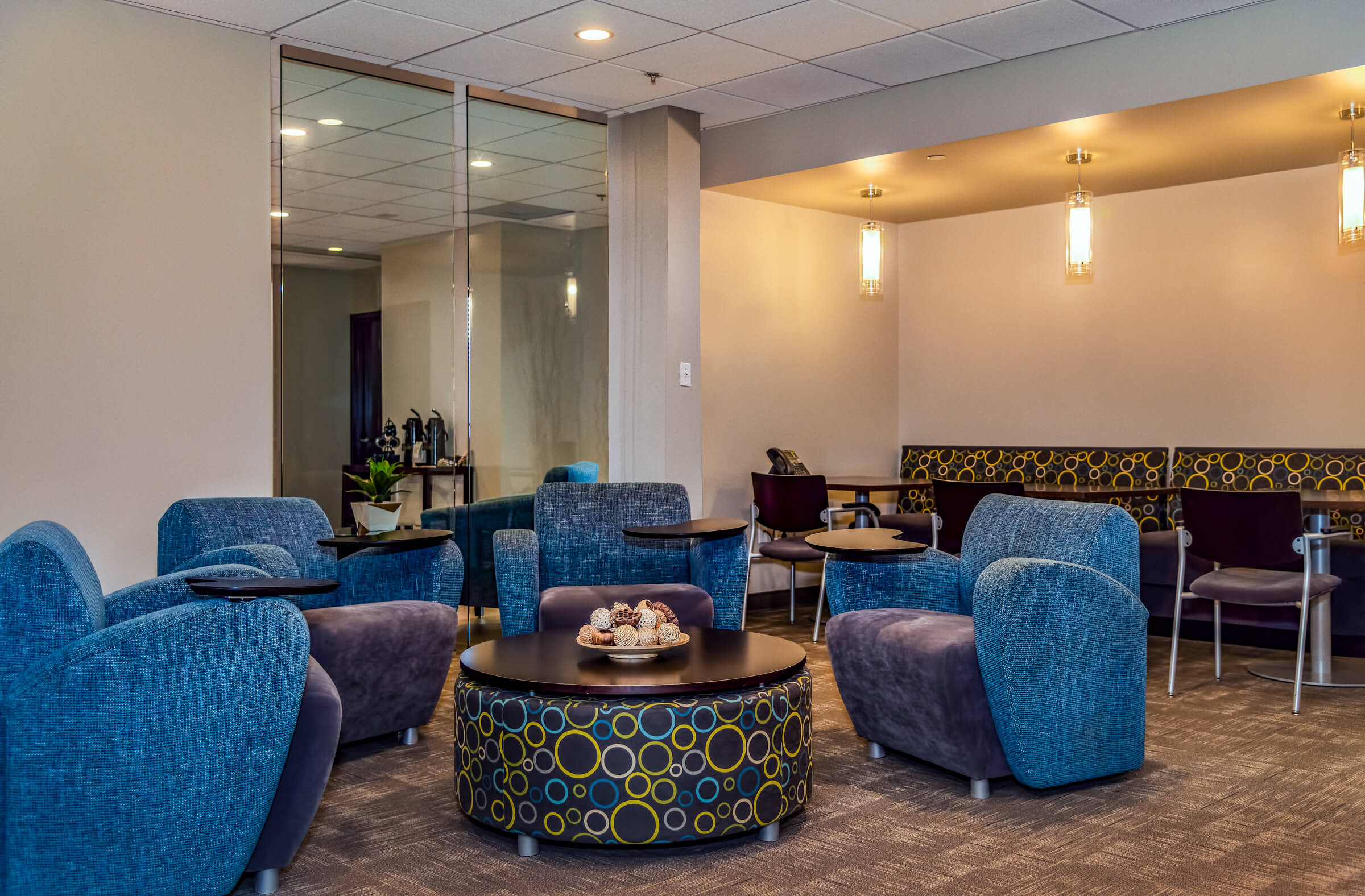 A modern, vibrant office lounge area with comfortable blue and purple armchairs, a patterned circular coffee table holding snacks, and an adjacent casual dining area with built-in seating.