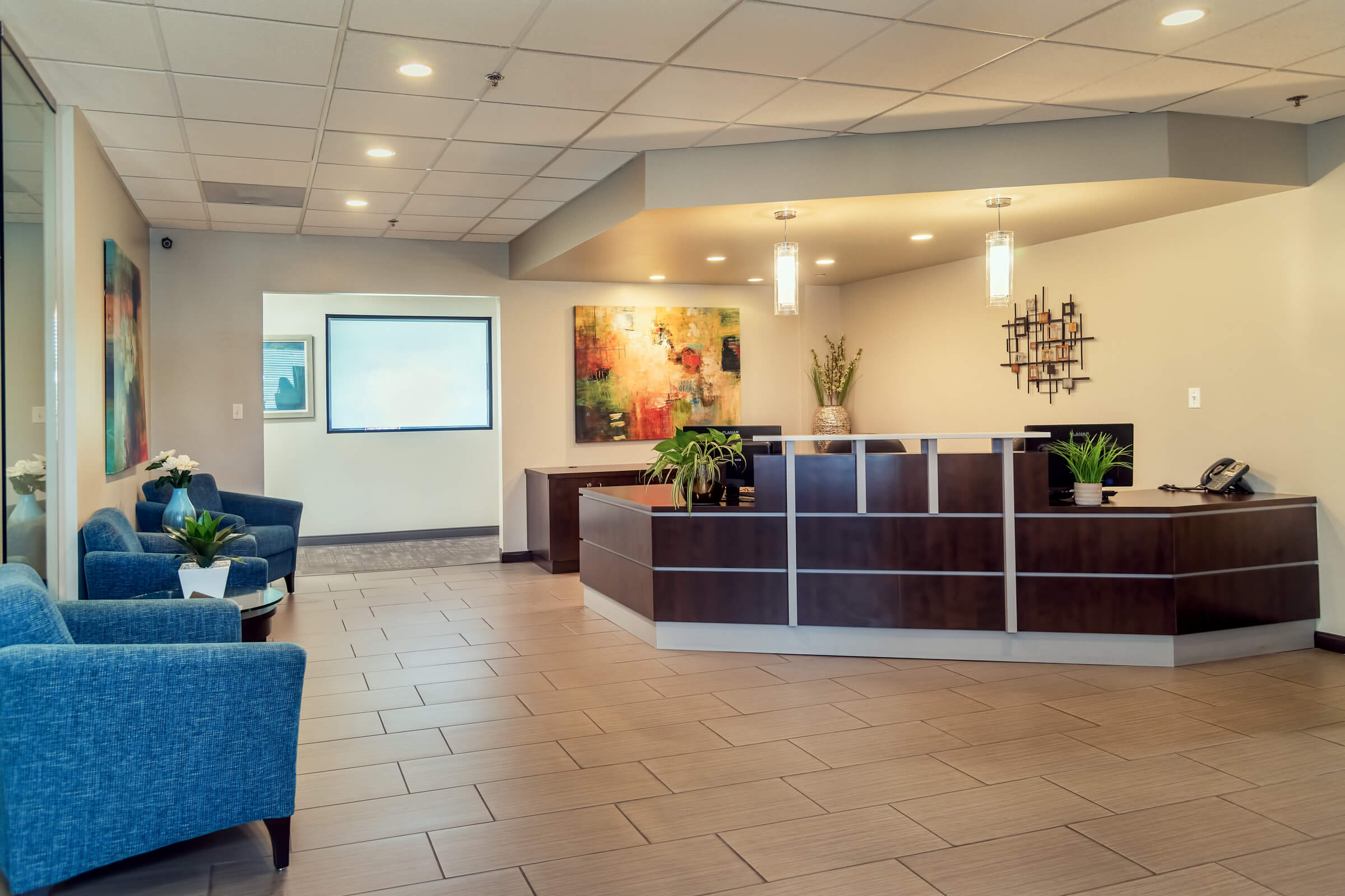 A welcoming, modern corporate reception area featuring a dark wood front desk, light beige tile flooring, a blue-chair seating area, and two large pieces of contemporary abstract art.