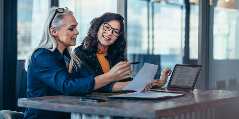Two,Women,Analyzing,Documents,While,Sitting,On,A,Table,In