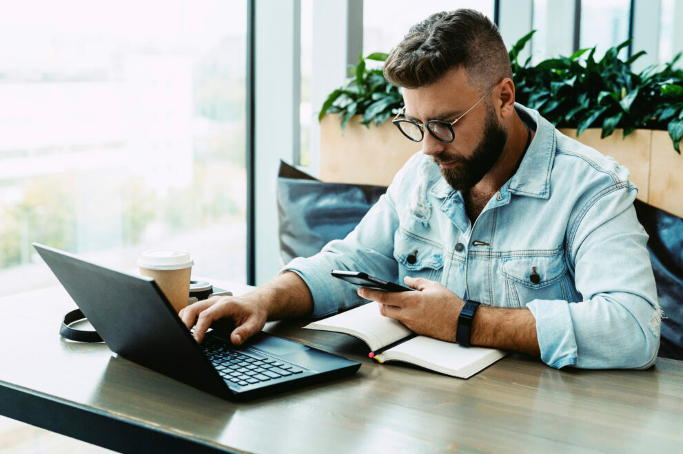 Hipster,Man,Sits,In,Cafe,,Uses,Smartphone,,Works,On,Laptop.