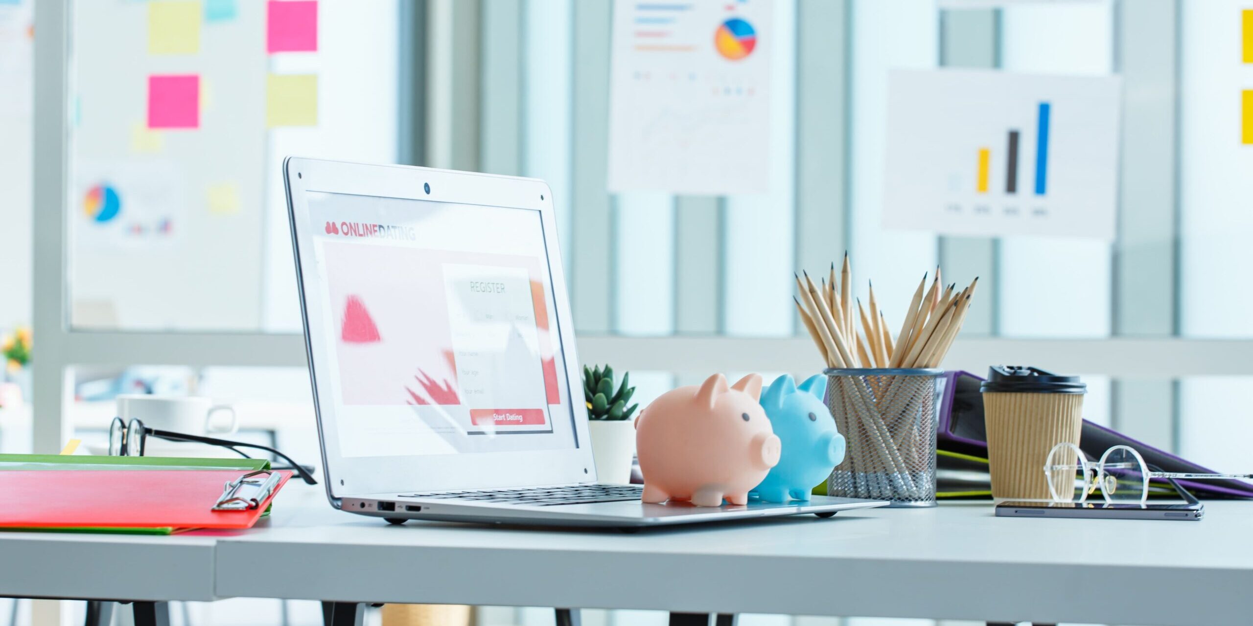 Financial Planning Workspace with Visual Data and Savings Tools White desk with laptop, piggy banks, coffee cup, pencils, and financial charts in a bright office setting.