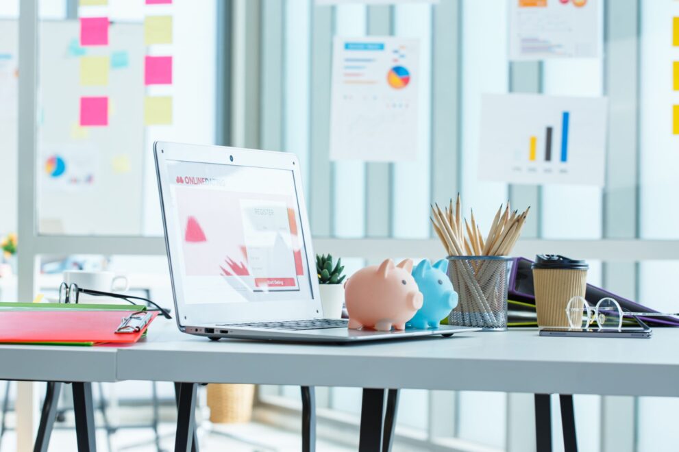 White desk with laptop, piggy banks, coffee cup, pencils, and financial charts in a bright office setting.